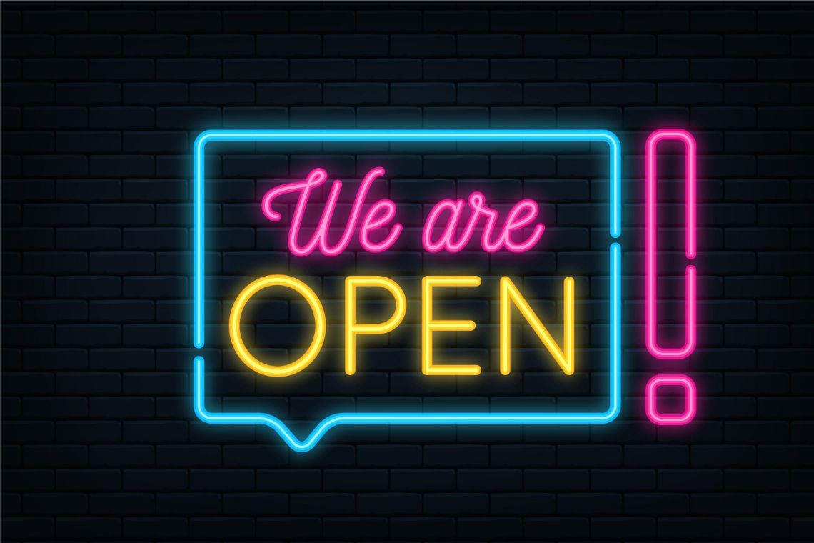 A neon sign displaying 'We are OPEN!' on a black brick background. PW Shop.